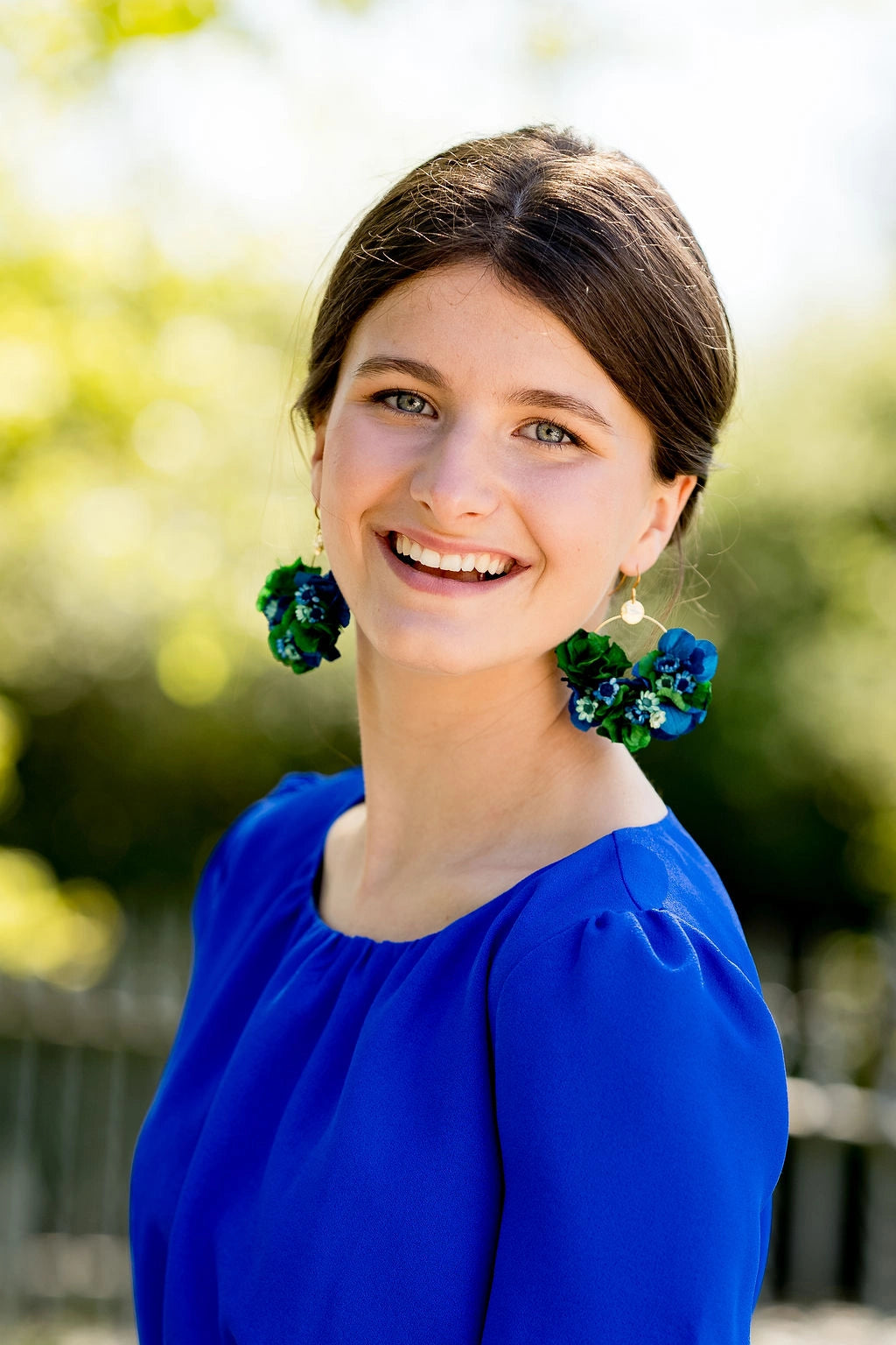 Jeune femme habillée d'une robe bleue roi avec des boucles d'oreilles en forme de créoles surmontés de fleurs d'hortensia et glixia séchées et stabilisées dans les tons verts et bleus. Créé par Bellemond.