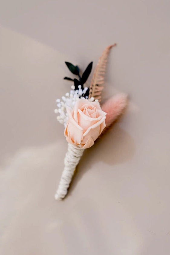 Boutonnière composée de fleurs séchées et stabilisées dans les tons rose, blanc et vert foncé. Créé par Bellemond.