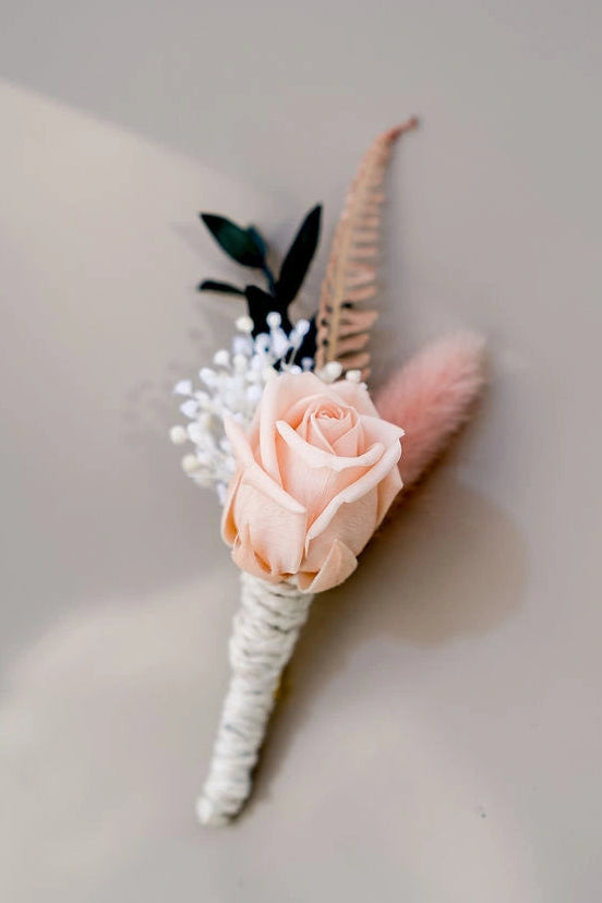 Boutonnière composée de fleurs séchées et stabilisées dans les tons roses, vert foncé et blanc. Créé par Bellemond.