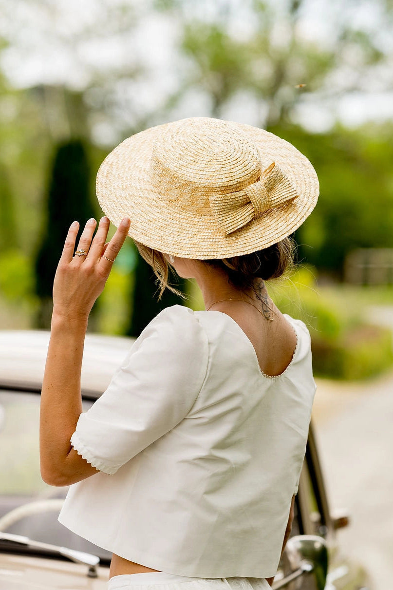 Femme se tenant de dos en haut de mariée, portant sur la tête un canotier en paille naturelle avec un noeud en paille naturelle sur l'arrière du chapeau. Créé par Bellemond.
