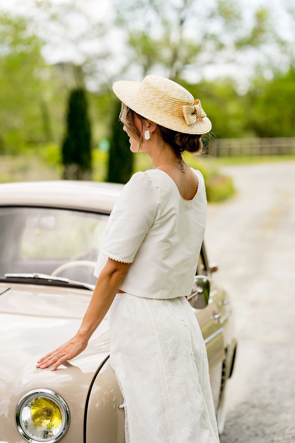 Mariée s'appuyant sur le capot d'une voiture portant sur la tête un canotier en paille naturelle avec un noeud en paille naturelle sur l'arrière du chapeau. Créé par Bellemond.