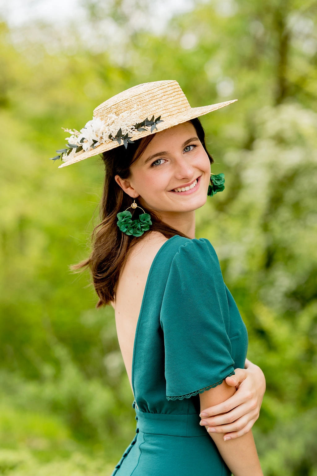 Jeune femme en robe verte portant sur la tête un canotier en paille naturelle surmonté d'une composition florale en fleurs séchées et stabilisées dans les tons verts, blancs et or. Créé par Bellemond.