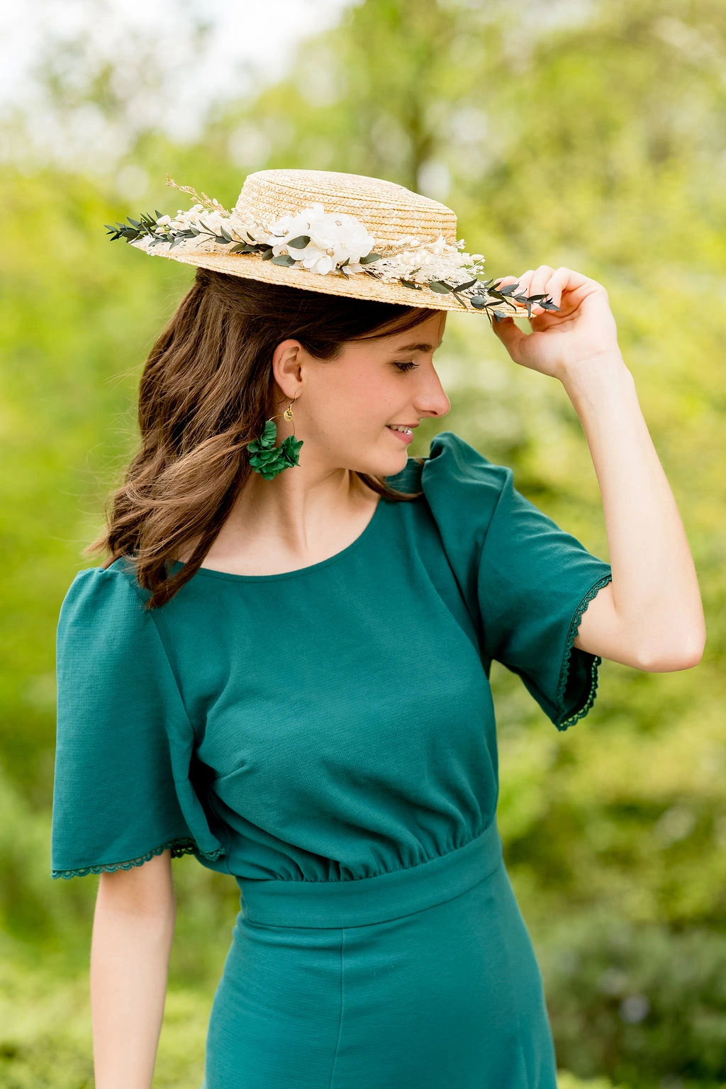 Jeune femme en robe verte portant sur la tête un canotier en paille naturelle surmonté d'une composition florale de fleurs séchées et stabilisées dans les tons verts, blancs et or. Créé par Bellemond.