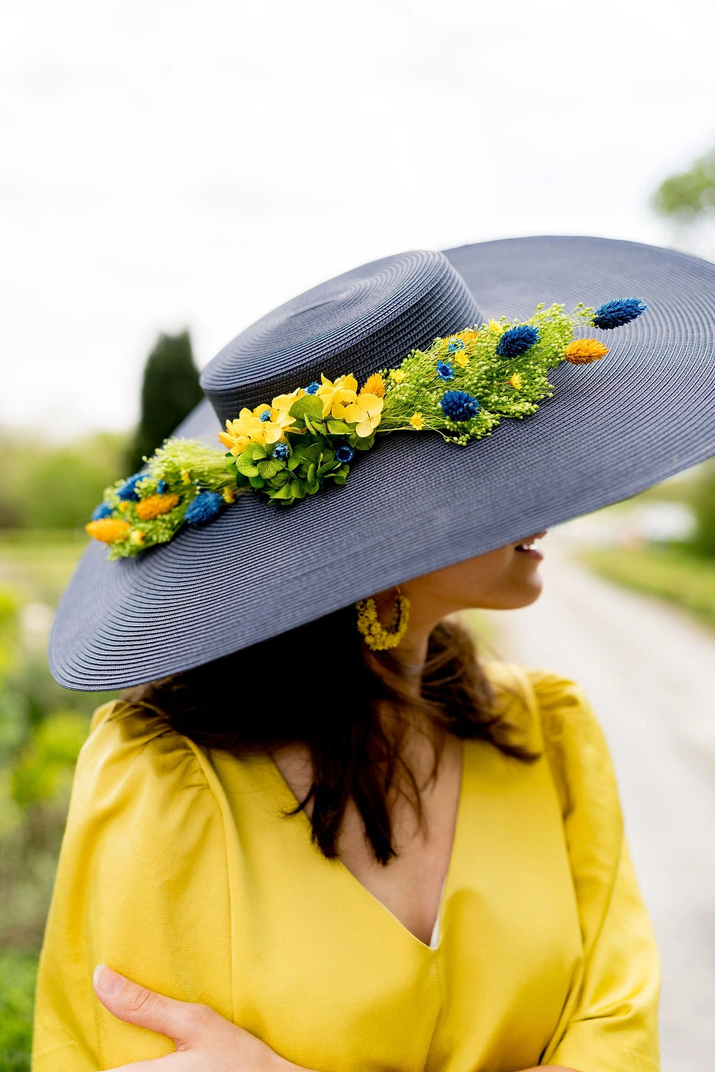 Jeune femme en robe jaune ayant sur la tête une grande capeline bleue marine avec une composition florale dans les tons bleus, jaunes et verts. Créé par Bellemond.