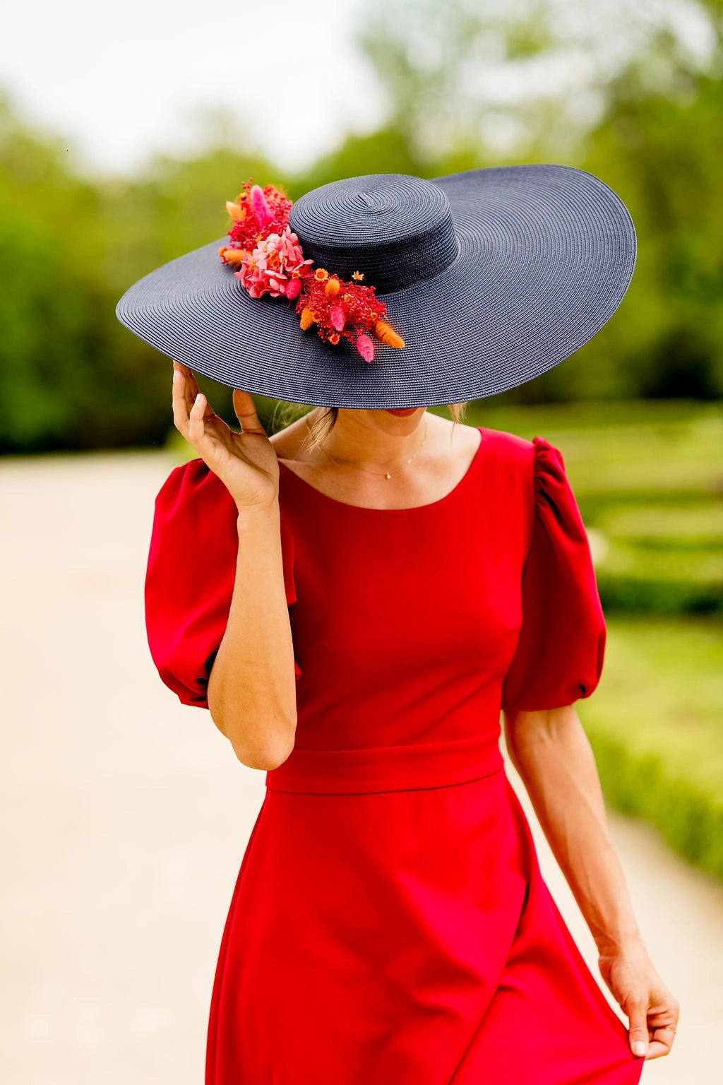 Jeune femme en robe rouge portant sur la tête une grande capeline bleue marine avec une composition florale en fleurs séchées dans les tons rouges, oranges et roses. Créé par Bellemond.