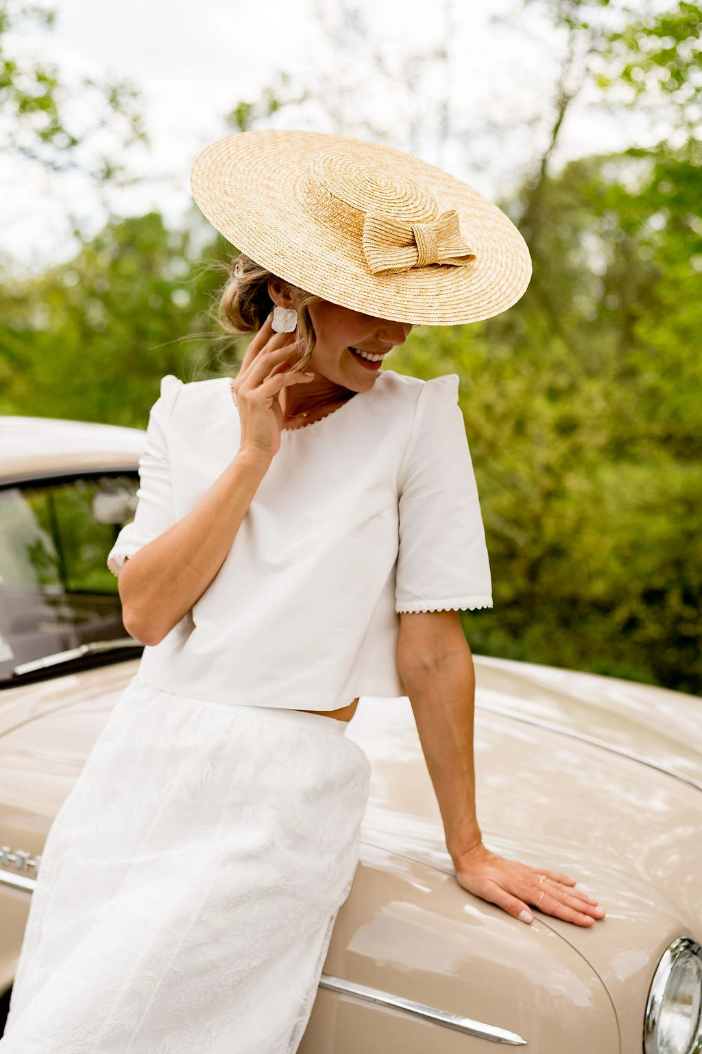 Jeune mariée s'appuyant sur le capot de la voiture portant sur la tête une capeline en paille surmonté d'un noeud en paille naturelle sur le devant. Créé par Bellemond.