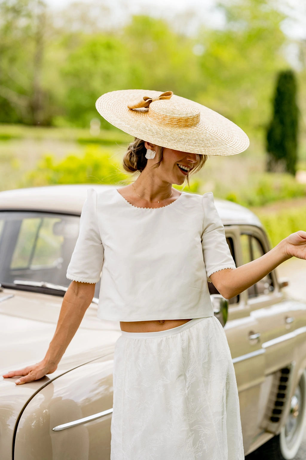 Jeune mariée se tenant contre une voiture et portant sur la tête une capeline en paille naturelle avec un noeud en paille naturelle à l'arrière. Créé par Bellemond.