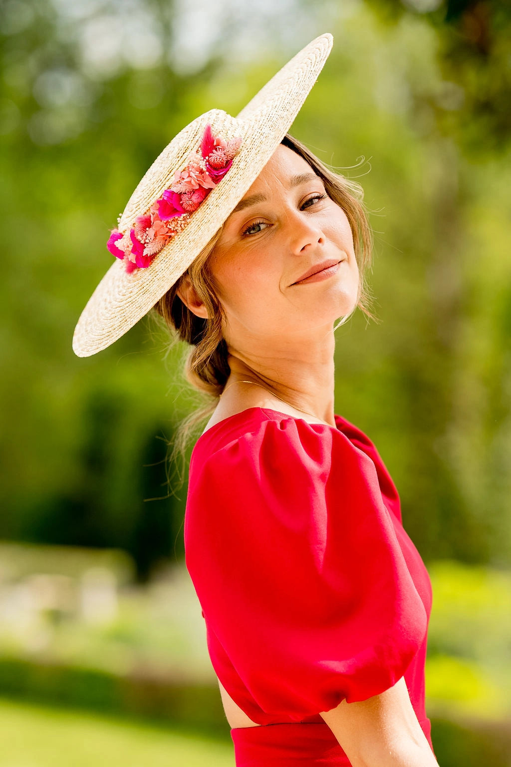 Jeune femme souriante portant sur la tête une capeline en paille naturelle sur laquelle sont montés des fleurs séchées et stabilisées autour de la calotte dans les tons fuchsia, corail et roses. Créé par Bellemond.