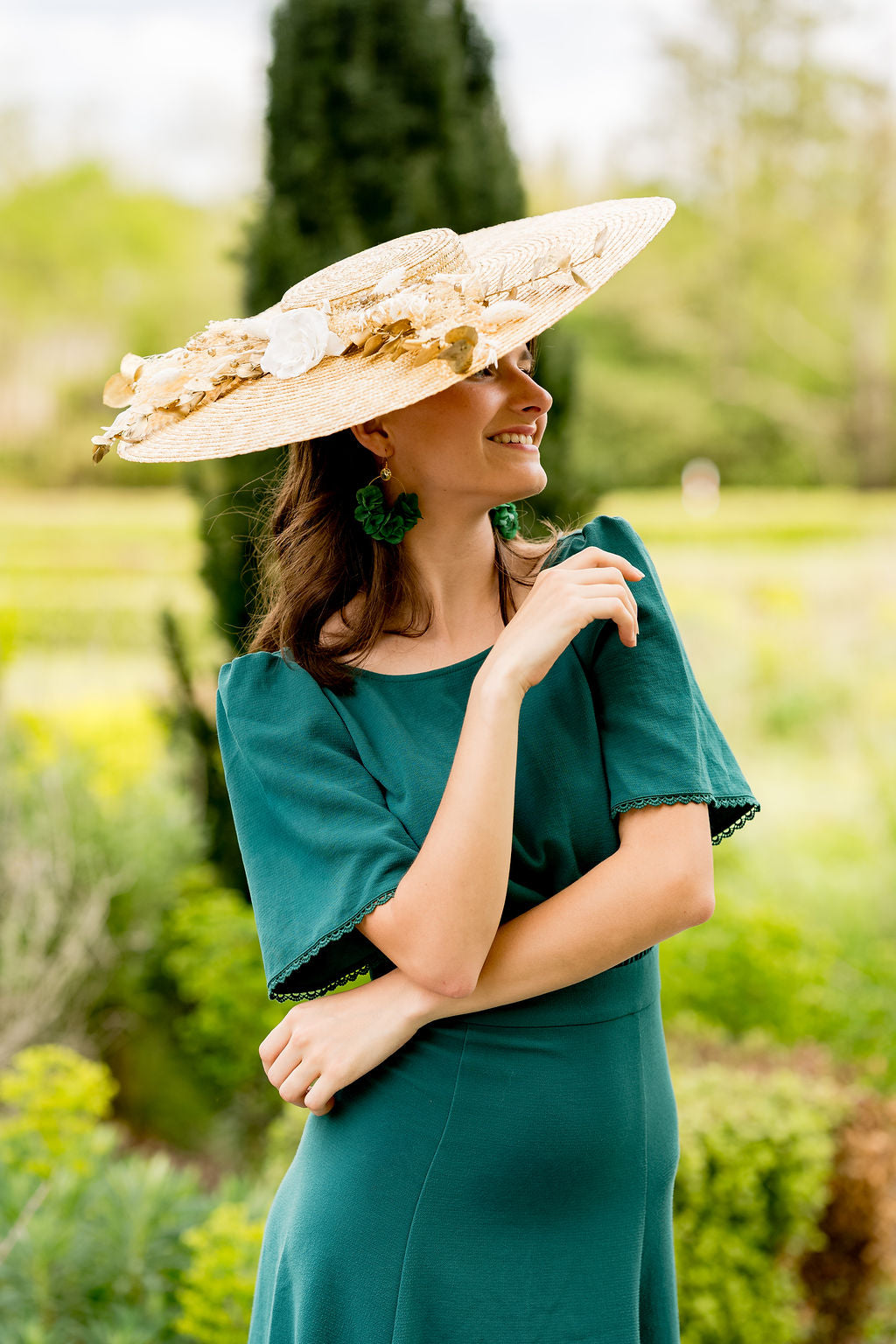 Jeune femme vêtue d'une robe verte ayant sur la tête une capeline en paille naturelle surmontée de fleurs séchées et stabilisées couleur blanc et or. Créé par Bellemond.