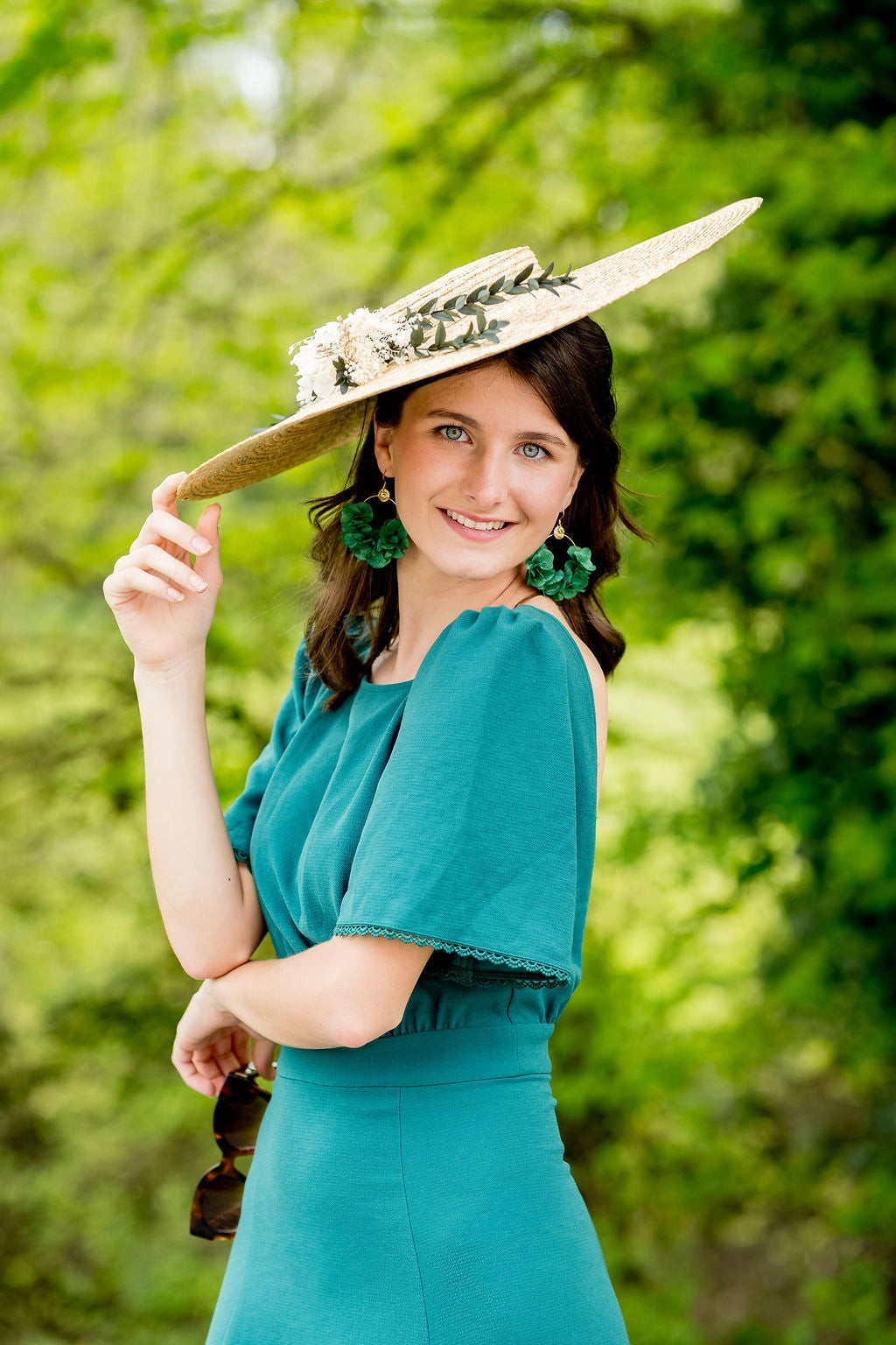 Jeune femme en robe verte portant sur la tête une grande capeline surmontée d'une composition florale blanche verte et dorée de fleurs séchées et stabilisées. Créé par Bellemond.