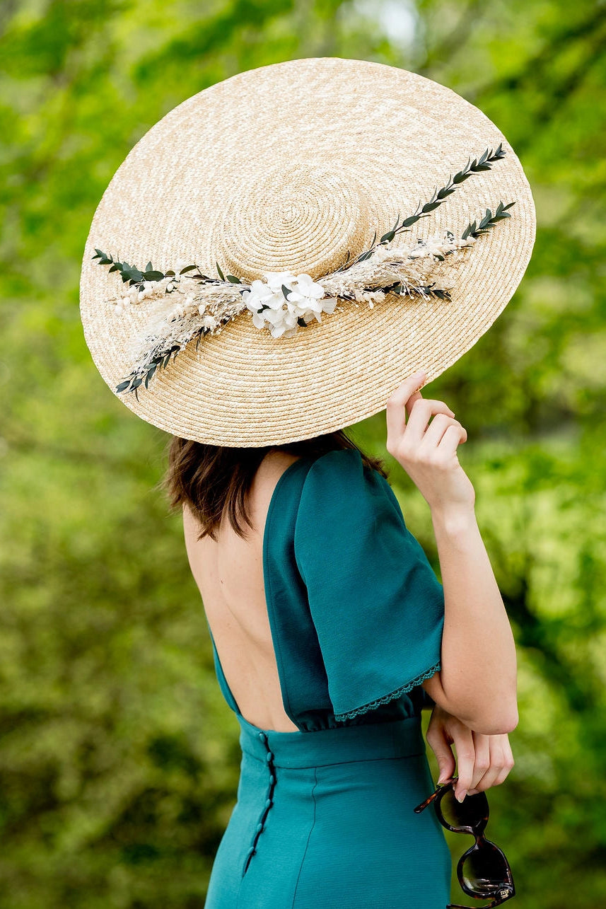 Jeune femme en robe verte se tenant de dos et portant sur la tête une grande capeline de paille naturelle surmonté de fleurs séchées et stabilisées dans les tons verts, blancs et dorés. Créé par Bellemond.