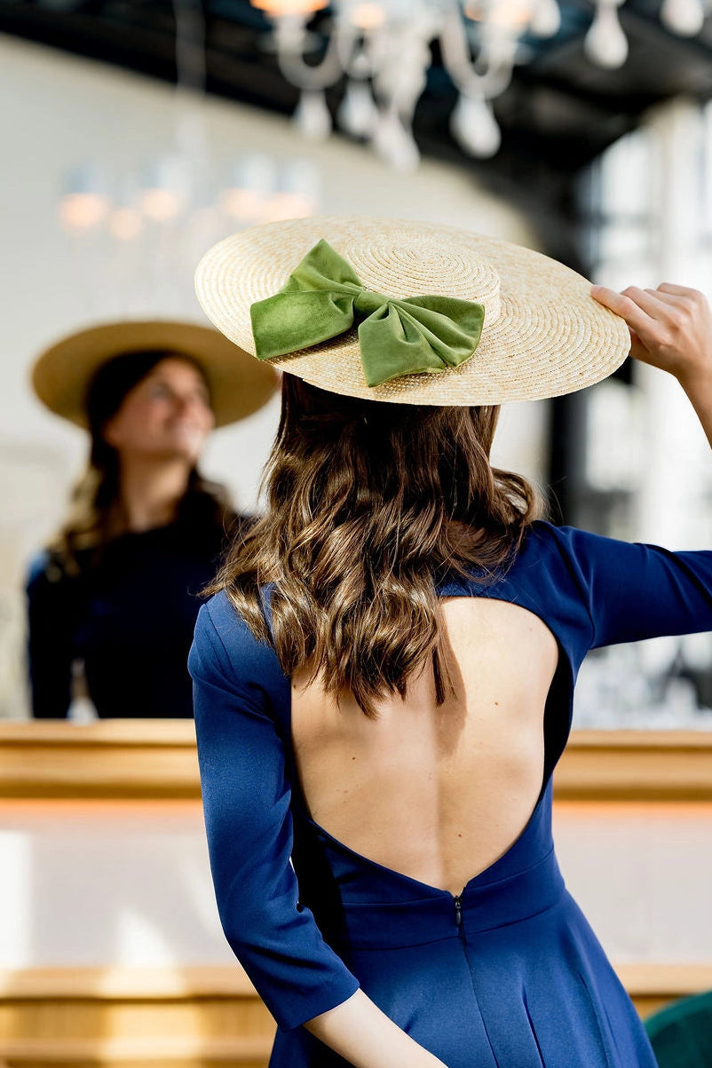 Jeune femme en robe bleue marine avec une capeline en paille sur la tête avec un noeud en velours vert olive. Créé par Bellemond.