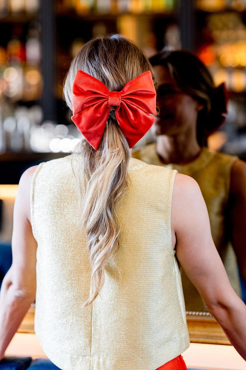 Jeune femme se tenant de dos avec un top doré et dans les cheveux une barrette avec un gros noeud en velours de couleur orange.