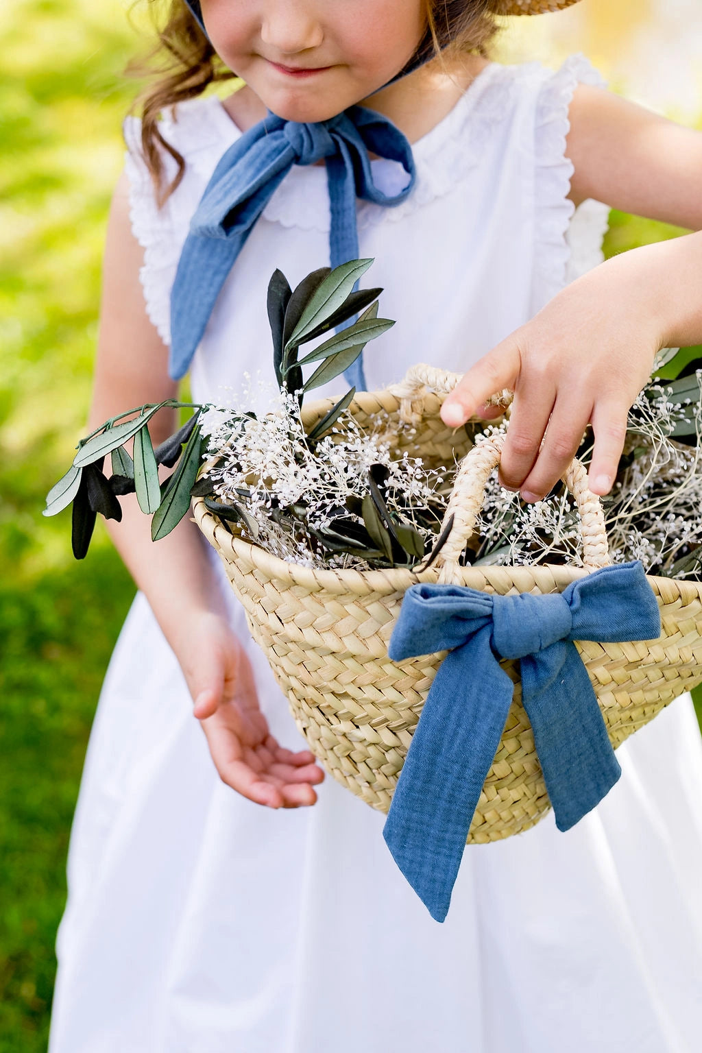 Petite fille de cortège montrant un petit panier tressé en feuilles de palmier sur lequel un noeud bleu gris est attaché avec une barrette. Créé par Bellemond.