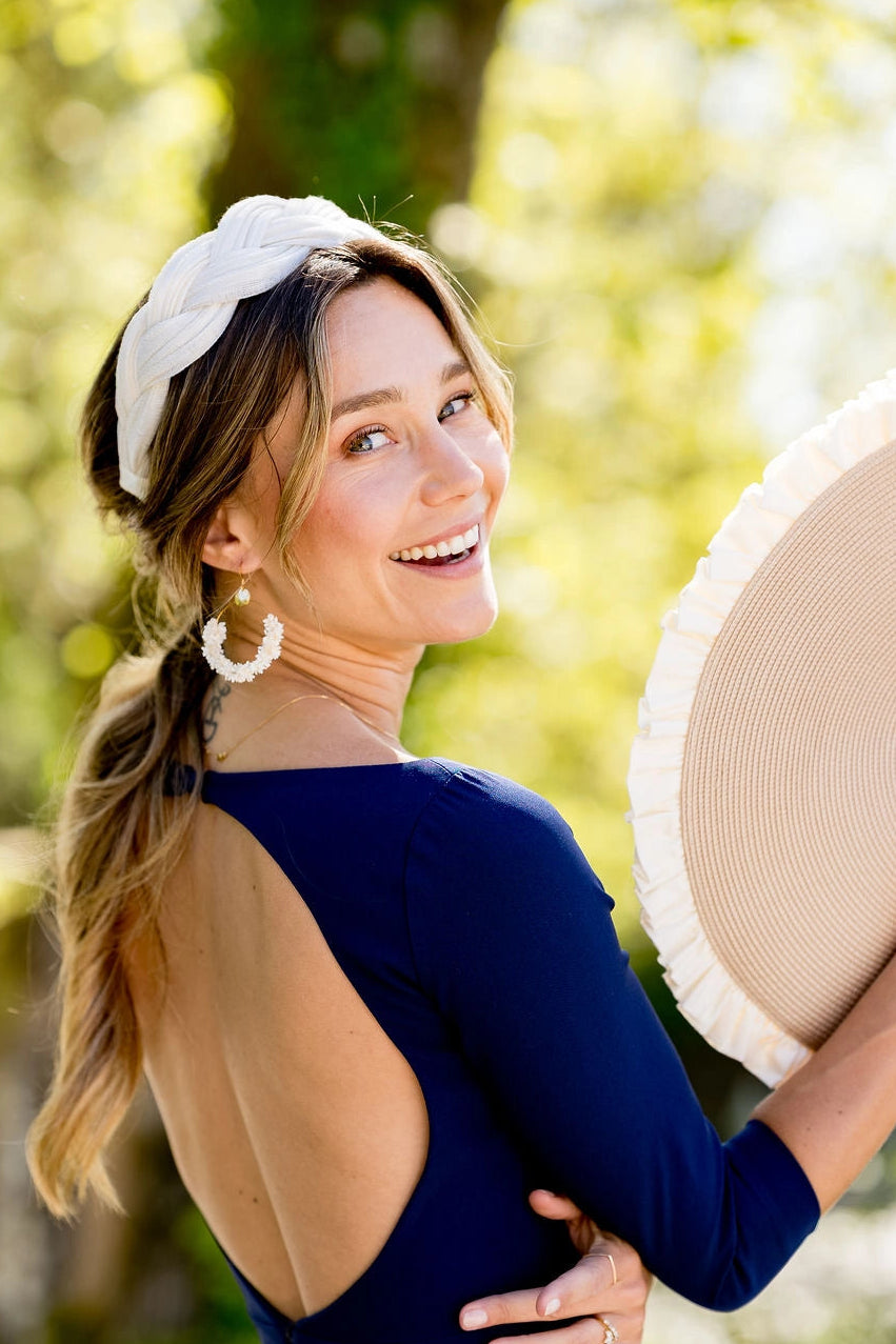 Jeune femme dans une robe bleue marine avec un serre-tête en sinamay tressé blanc ivoire et ayant à la main une pochette avec volants. Créé par Bellemond.