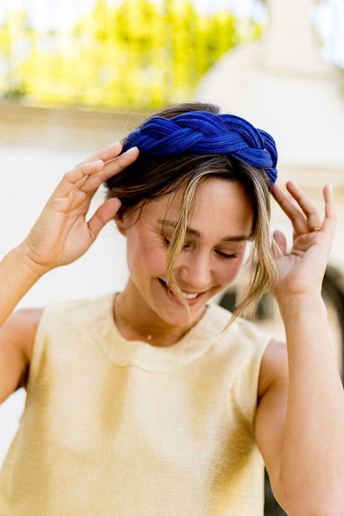 Jeune femme portant à la tête ses mains pour réajuster un serre-tête tressé en sinamay de couleur bleu. Créé par Bellemond.