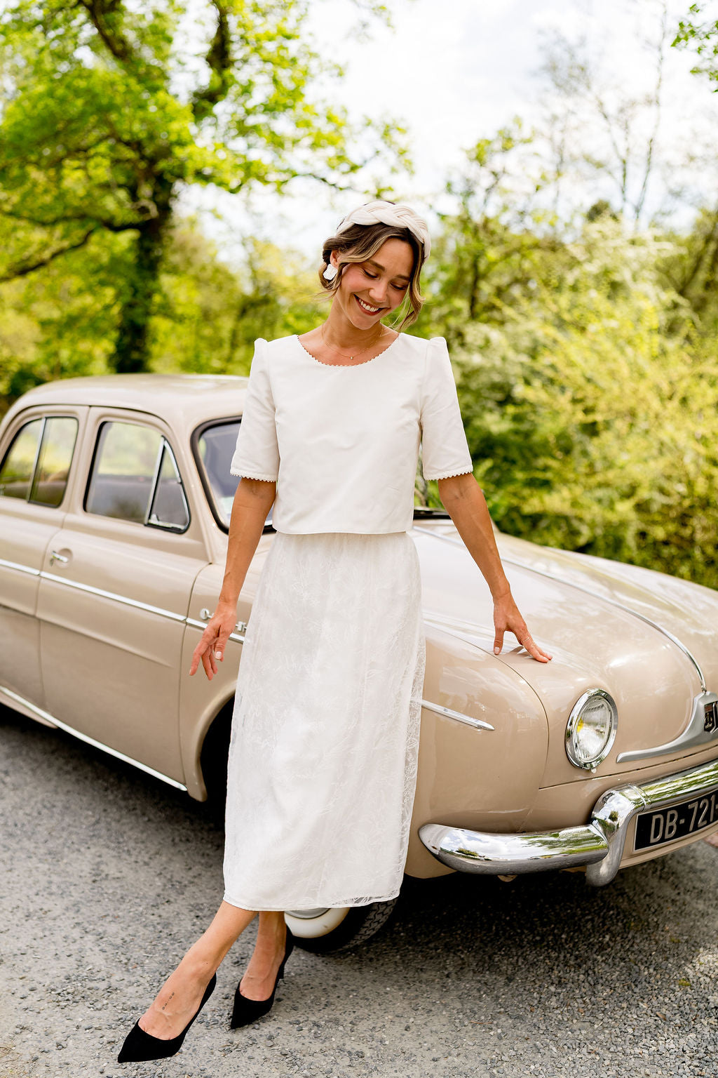 Une mariée devant une voiture vintage portant dans les cheveux un serre-tête tressé en sinamay de couleur ivoire. Créé par Bellemond.