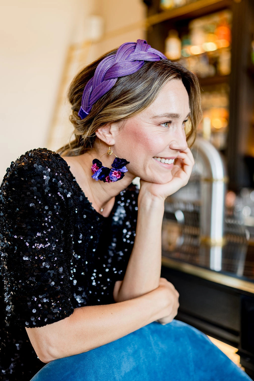 Jeune femme habillée d'un top à sequins noirs, portant un serre-tête tressé en sinamay de couleur violet ainsi que des boucles d'oreilles dans les tons violets également. Créé par Bellemond.