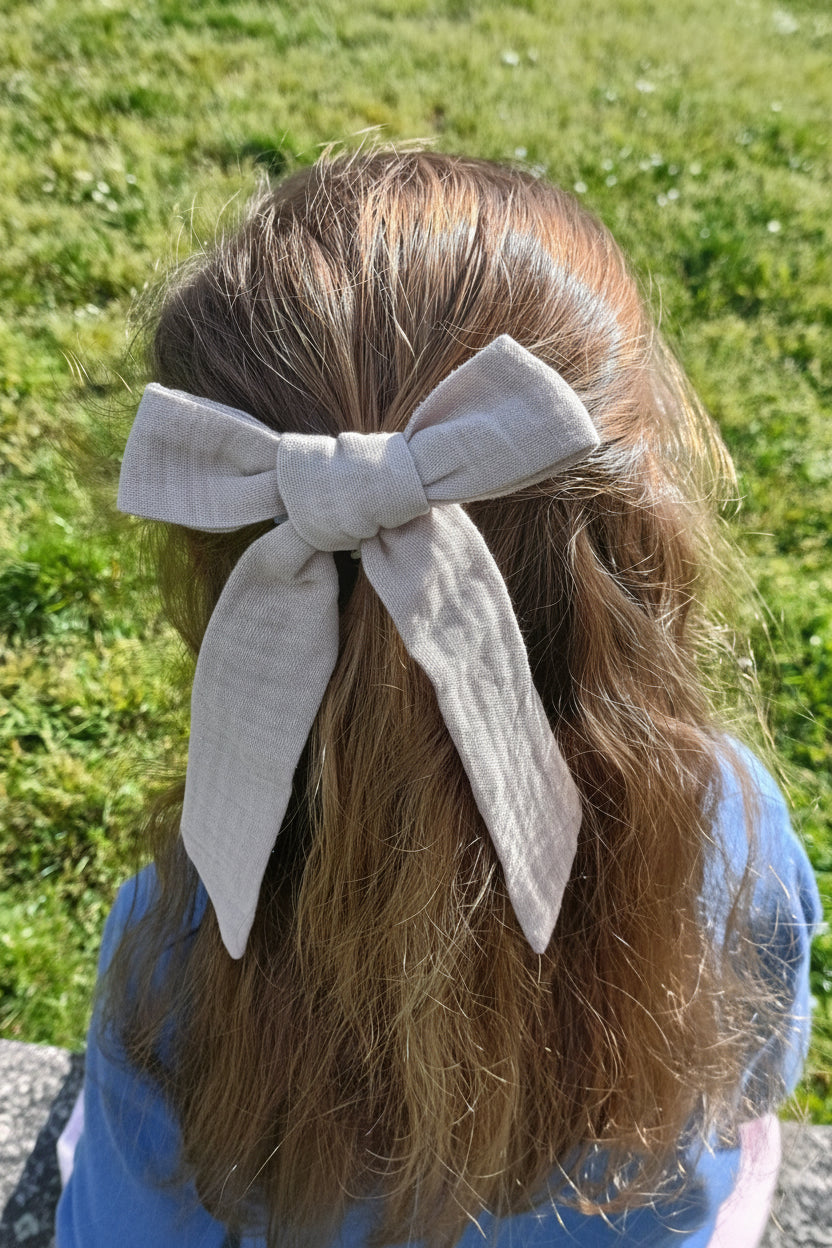 Petite fille se tenant de dos avec cheveux lâchés dans lesquels se trouve une barrette en gaze de coton de couleur taupe. Créé par Bellemond.
