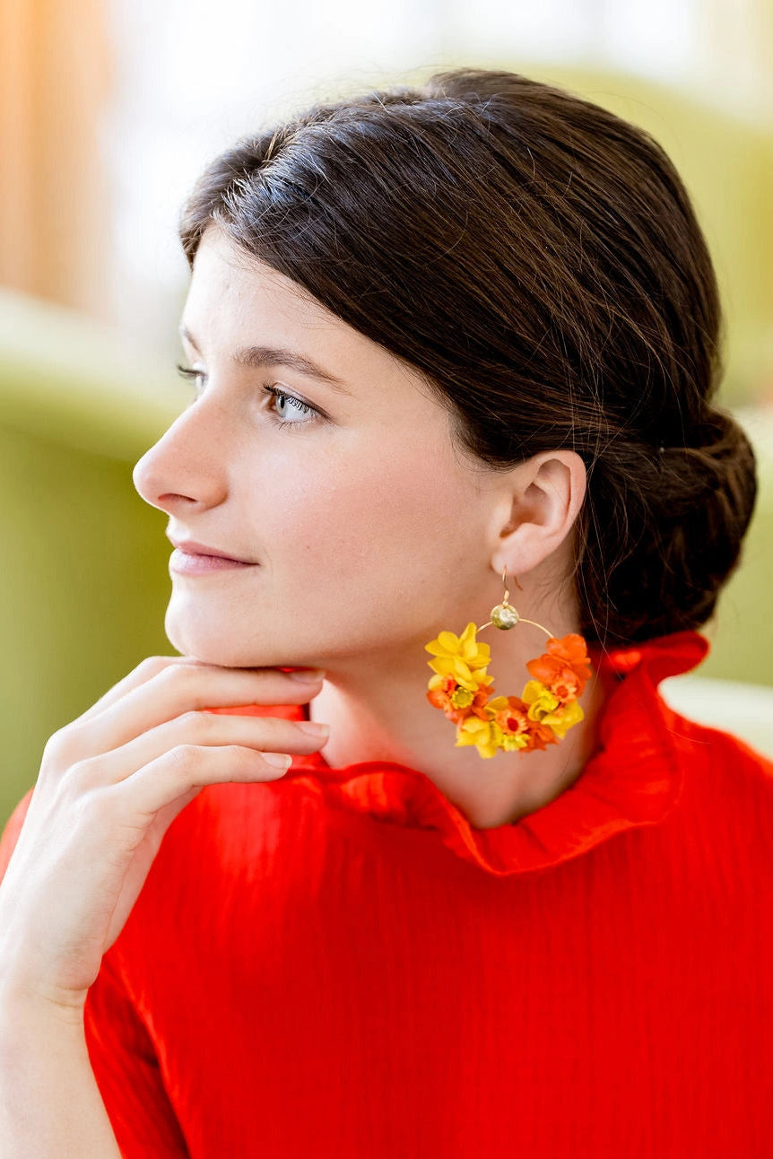 Jeune femme avec haut rouge ayant des boucles d'oreilles en forme de créoles avec des fleurs séchées et stabilisées : hortensia et glixa de couleur orange et jaune. Créé par Bellemond.