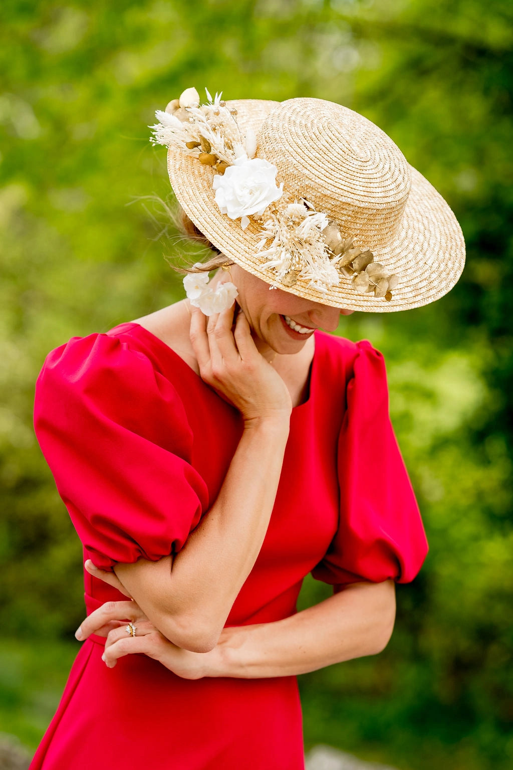 Jeune femme en robe rouge se penchant en avant ayant sur la tête un canotier en paille naturelle surmonté de fleurs séchées et stabilisées blanches et dorées. Créé par Bellemond. 