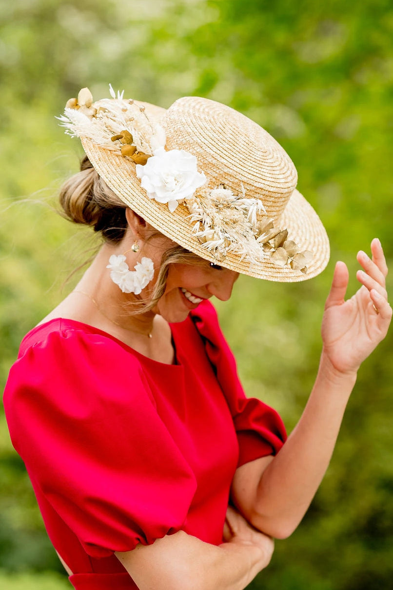 Jeune femme en robe rouge ayant sur la tête un canotier en paille naturelle surmonté de fleurs séchées et stabilisées dans les tons blancs et dorés. Crée par Bellemond. 