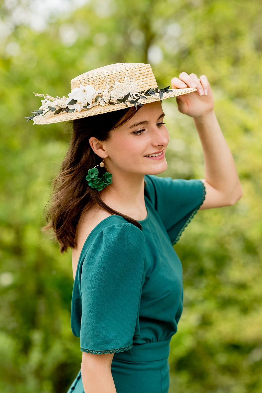 Jeune femme en robe verte et dans un cadre verdoyant portant sur la tête un canotier en paille naturelle surmonté d'une composition florale en fleurs séchées et stabilisées de couleurs vert, blanc et or. Créé par Bellemond.