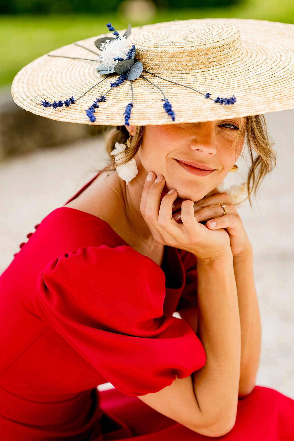 Jeune femme souriante en robe rouge tenant sur la tête une capeline en paille naturelle surmontée de lavande, mini gypsophile et eucalyptus. Créé par Bellemond. 