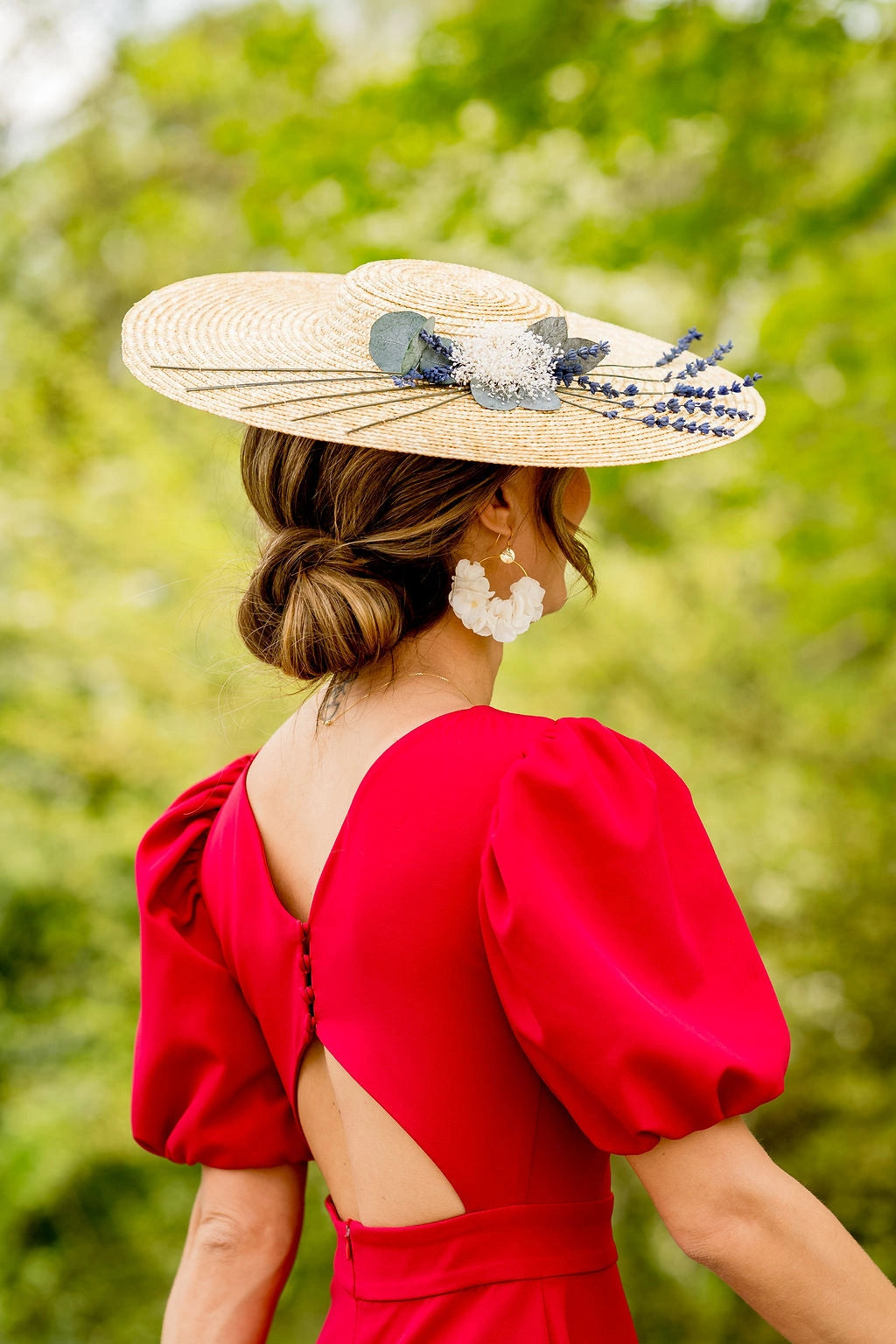 Jeune femme habillée d'une robe rouge tenant sur la tête une capeline en paille naturelle surmontée de lavande, d'eucalyptus et de mini-gypsophile. Créé par Bellemond.
