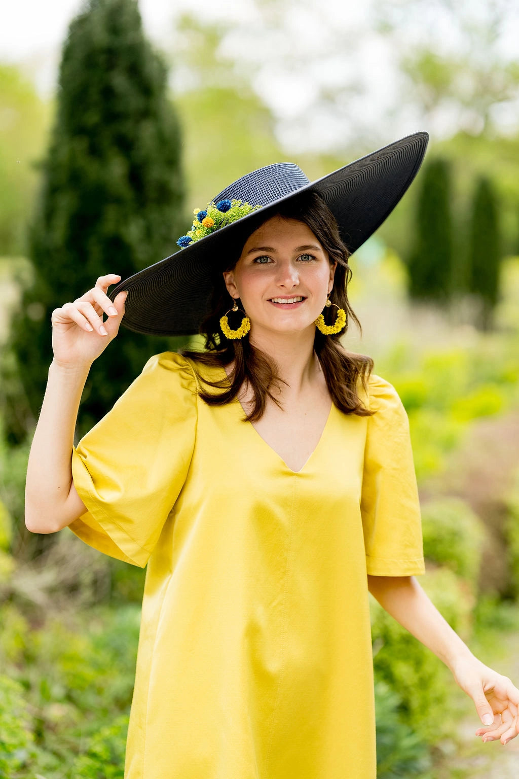 Jeune femme portant une robe jaune avec une grande capeline bleue marine sur la tête avec une composition florale dans les tons verts, jaunes et bleus. Créé par Bellemond. 