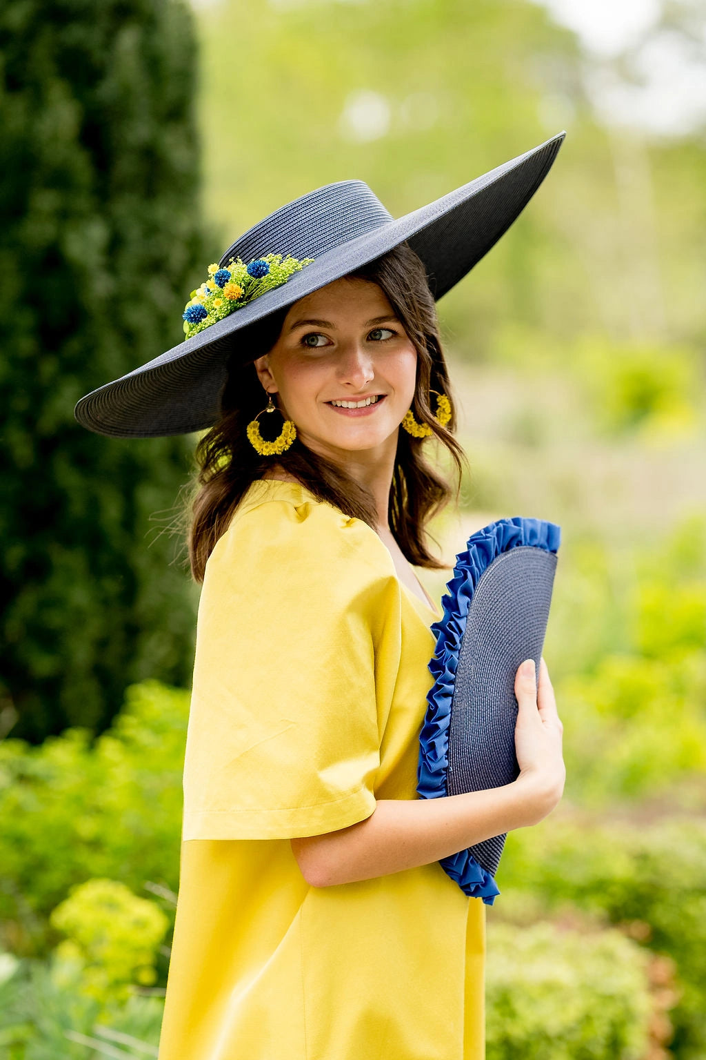 Jeune femme en robe jaune et tenant une pochette bleue marine et ayant sur la tête une grande capeline bleue marine avec une composition florale en fleurs séchées vertes, jaunes et bleues. Créé par Bellemond. 