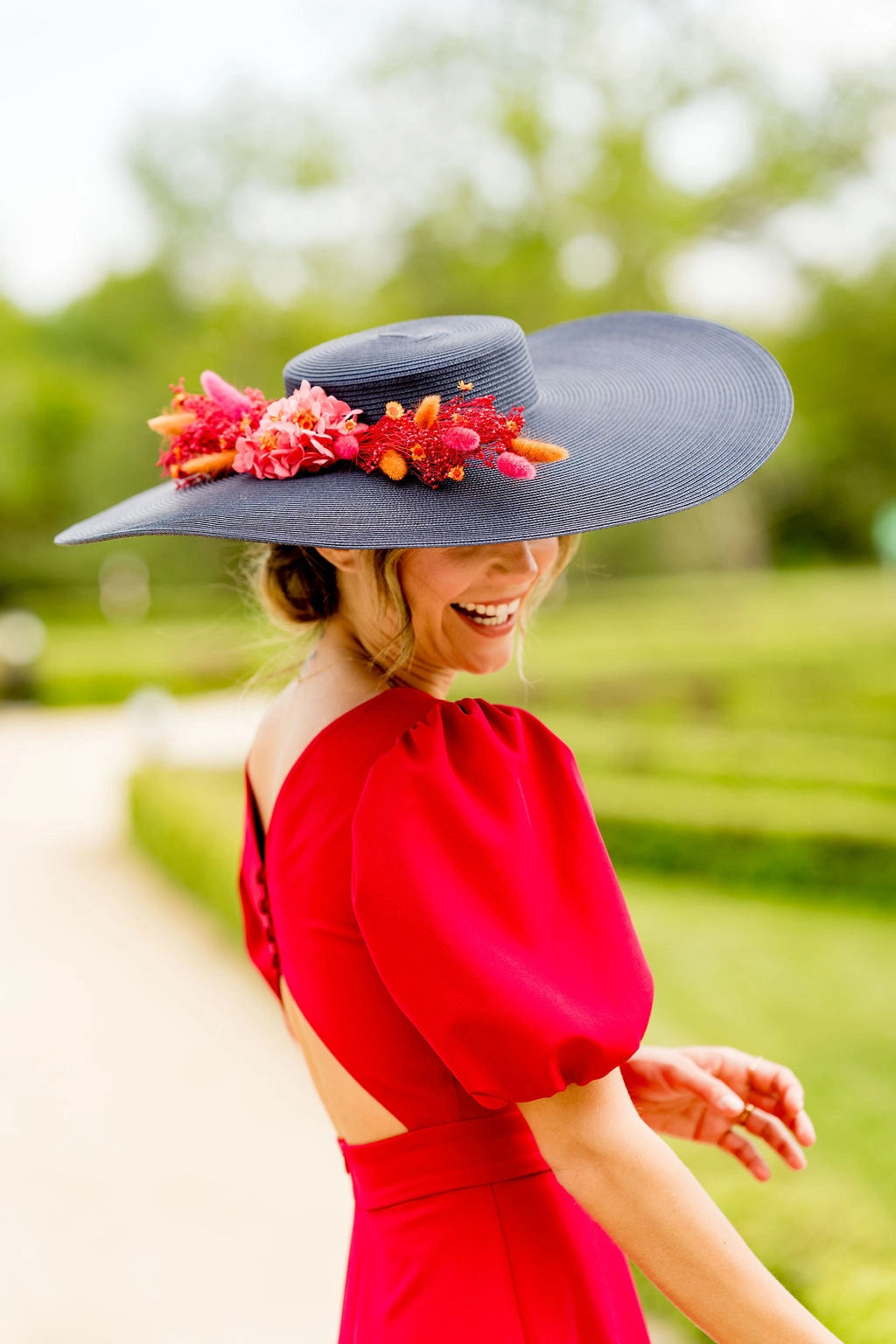 Jeune femme en robe rouge tenant sur la tête une capeline bleue marine avec une composition florale en fleurs séchées dans les tons oranges, rouges et roses. Crée par Bellemond. 