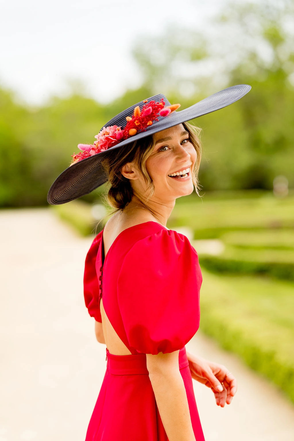 Jeune femme souriante avec une robe rouge dans un cadre verdoyant, ayant sur la tête une grande capeline bleue marine avec une composition florale en fleurs séchées et stabilisées dans les tons oranges, rouges et roses. Créé par Bellemond. 