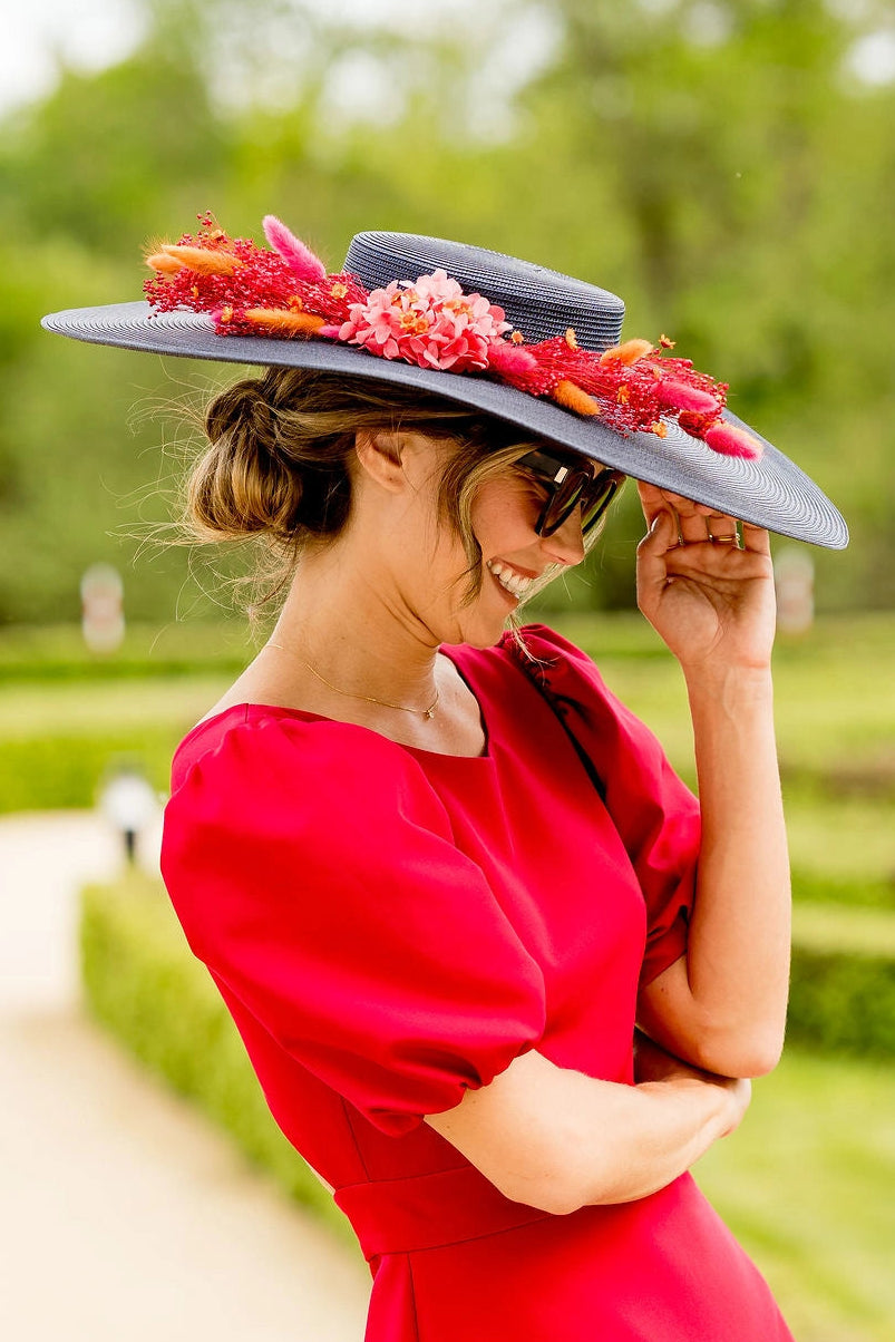 Jeune femme habillée d'une robe rouge tenant sur la tête une capeline bleue marine avec une composition florale dans les tons roses, rouges et oranges. Créé par Bellemond. 