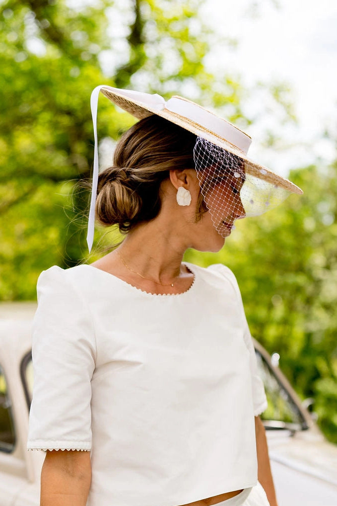 Jeune mariée se tenant proche d'une voiture ancienne portant sur la tête une capeline entouré d'un ruban en satin blanc et avec une voilette sur le devant du chapeau. Créé par Bellemond.