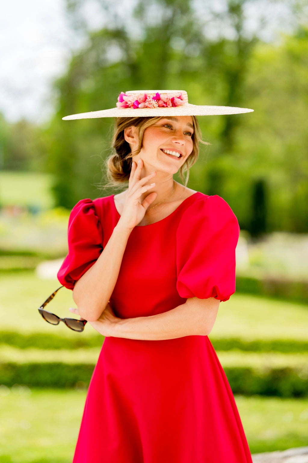 Jeune femme dans une robe rouge portant une grande capeline en paille naturelle surmonté de fleurs stabilisées et séchées de couleurs roses, fuchsia et corail. Créé par Bellemond.