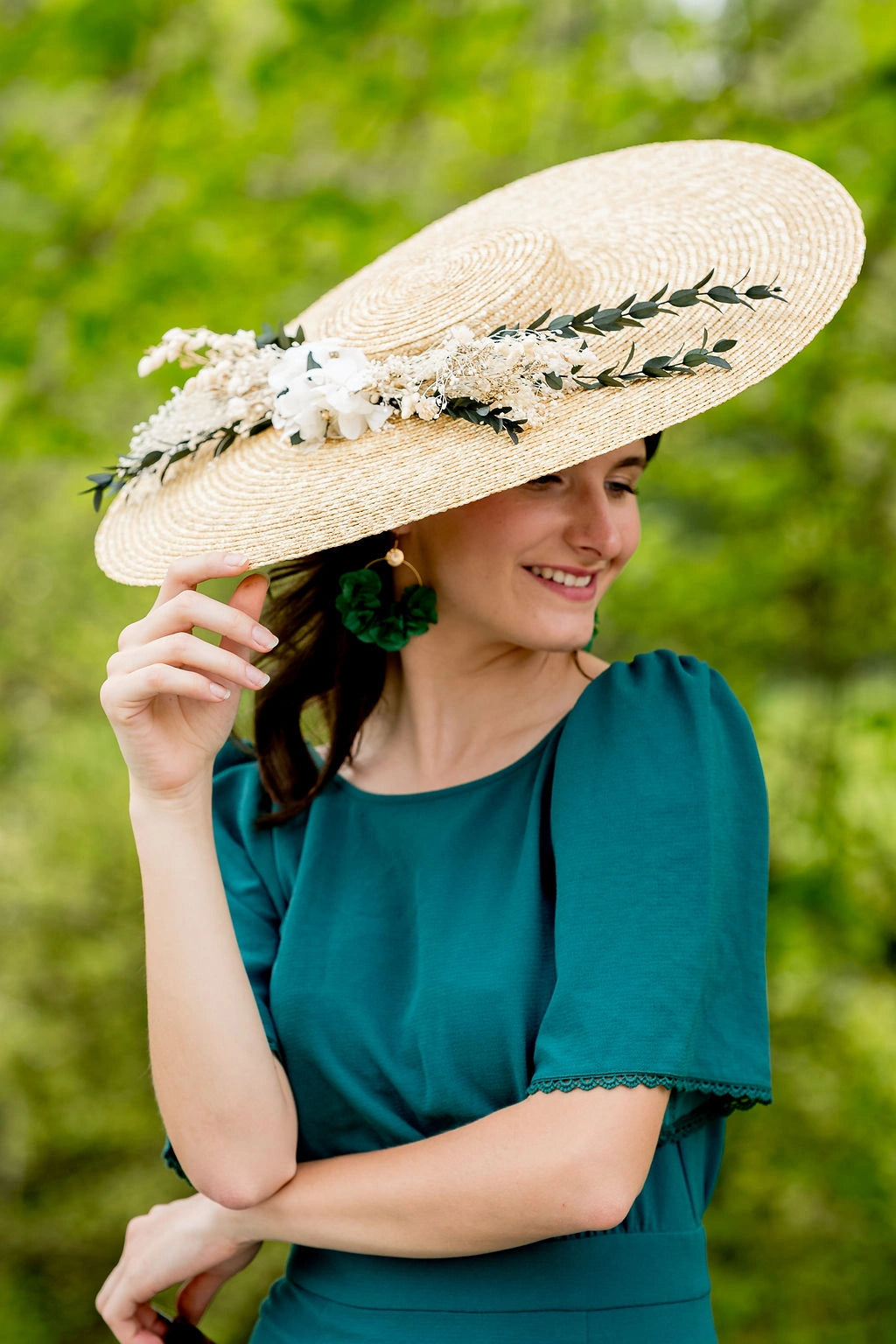 Jeune femme habillée d'une robe verte portant sur la tête une capeline surmontée d'une composition florale de fleurs séchées et stabilisées dans les tons blancs, or et verts. Créé par Bellemond. 