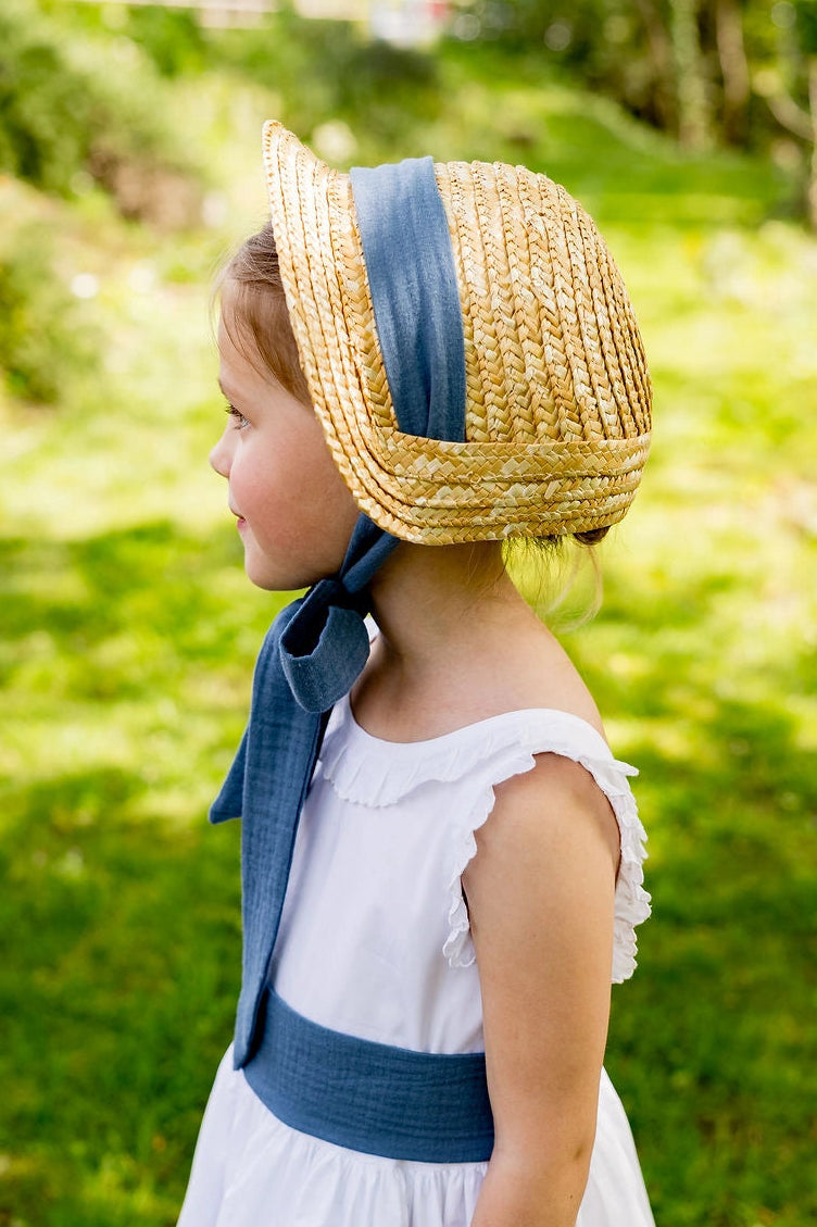 Petite fille demoiselle d'honneur portant un petit chapeau de type béguin en paille avec un ruban en gaze de coton bleu et gris. Créé par Bellemond. 