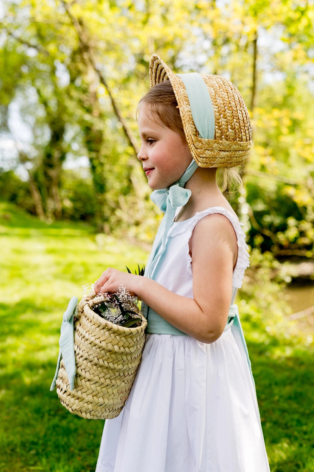 Petite fille portant un chapeau en paille de type calotte avec un ruban en gaze de coton de couleur vert menthe. Elle est dans un cadre verdoyant. Le chapeau est crée par Bellemond. 