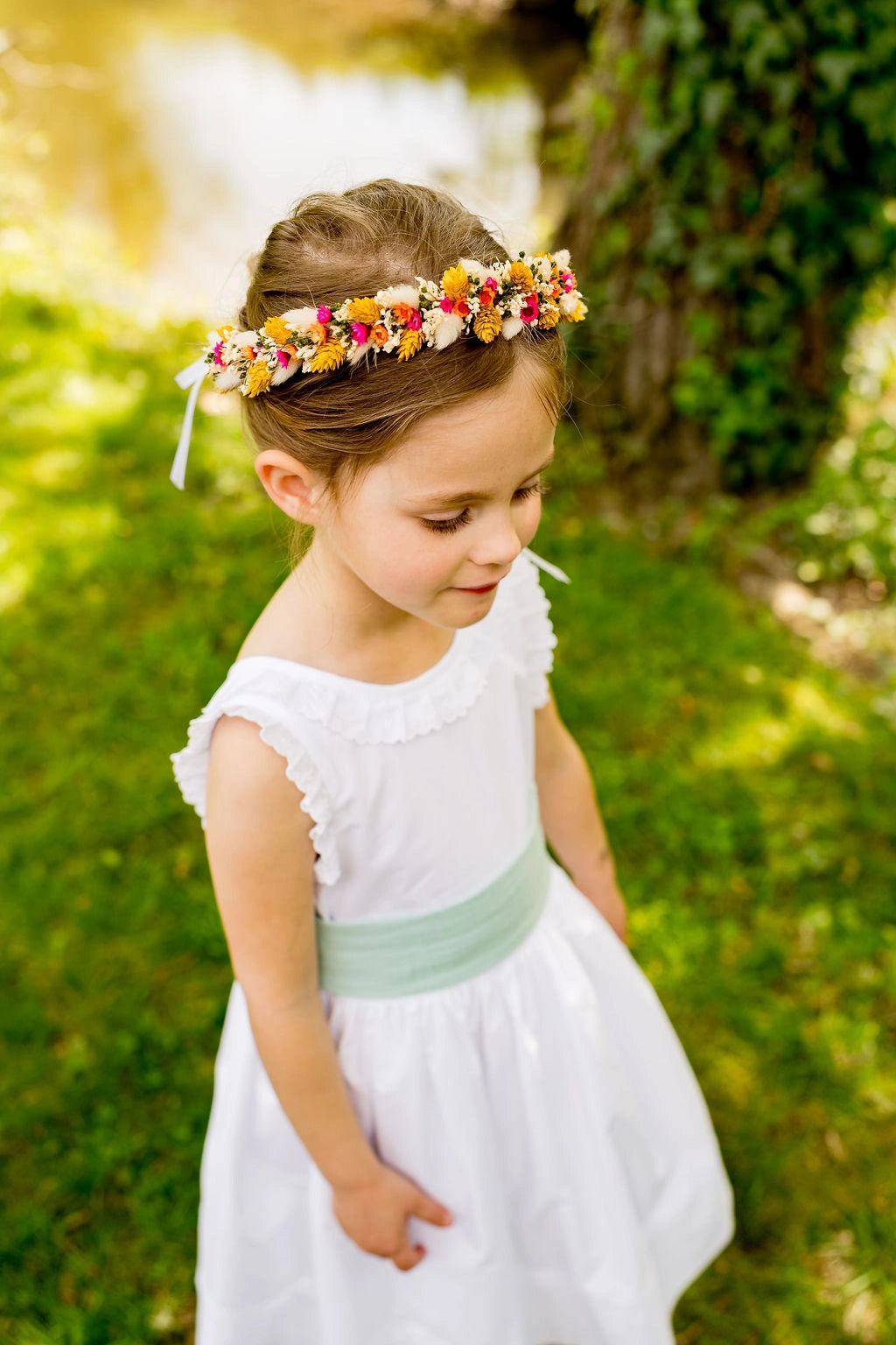Petite fille en robe blanche portant une couronne de fleurs multicolore. Les fleurs sont stabilisées ou séchées. Créé par Bellemond. 