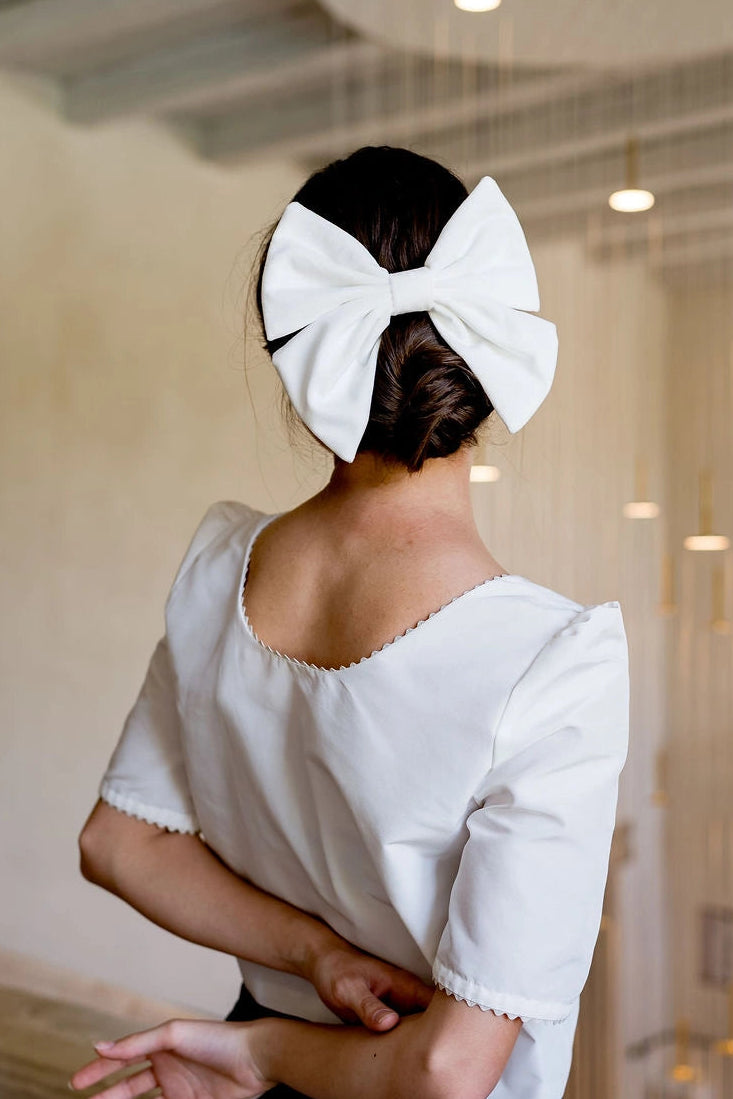 Jeune femme se tenant de dos avec un haut blanc ayant dans les cheveux un noeud en velours blanc monté sur barrette au dessus de son chignon. Créé par Bellemond. 