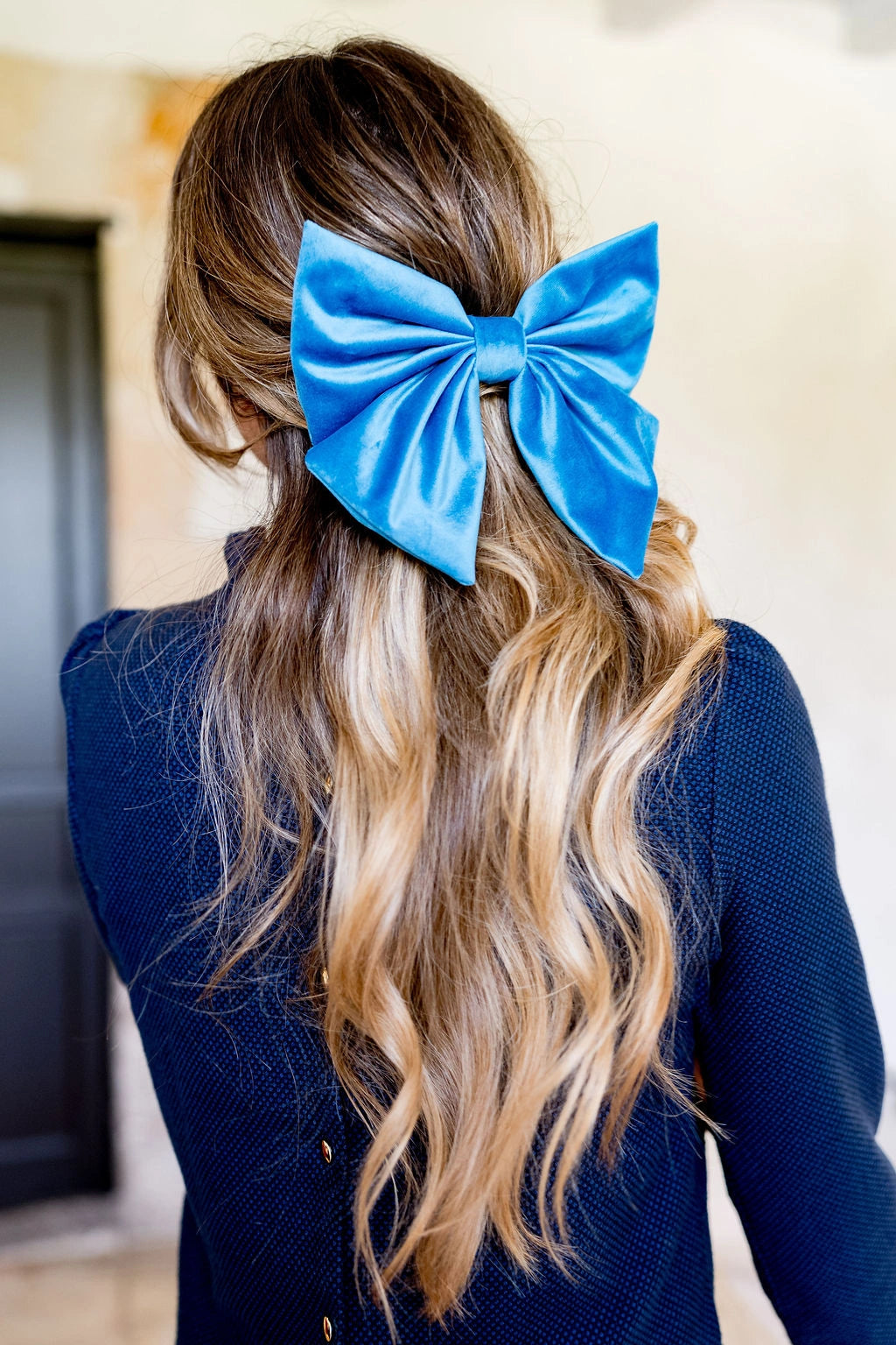 Jeune femme se tenant de dos avec un haut bleu marine et les cheveux lâchés dans lesquelles se trouve un noeud en velours de couleur bleu paon sous forme de barrette. Créé par Bellemond. 
