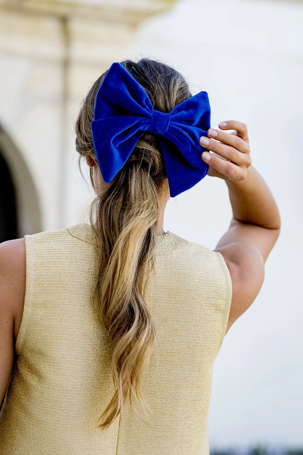Jeune femme se tenant de dos avec un top doré et touchant le noeud en velours dans sa chevelure qui est monté sur barrette et a une couleur bleue roi. Créé par Bellemond. 