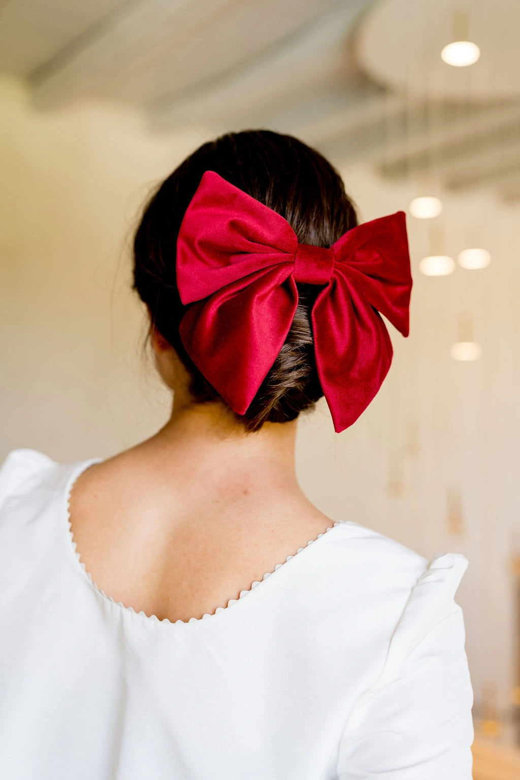 Jeune femme se tenant de dos habillée d'un haut blanc ayant dans les cheveux une barrette en forme de noeud en velours couleur rouge. Créé par Bellemond. 