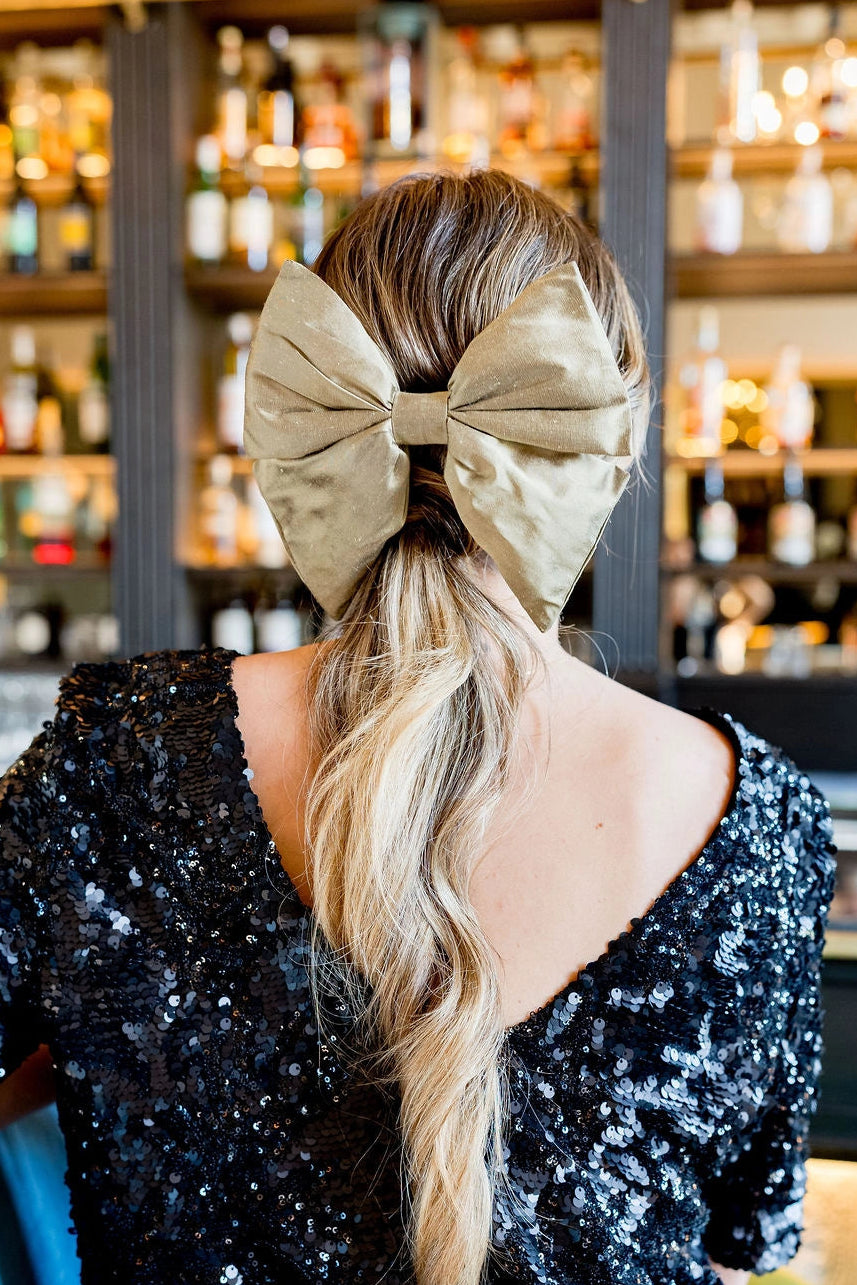 Jeune femme se tenant de dos assise dans un bar avec un top en sequins noirs ayant dans les cheveux une barrette sous forme de noeud en soie et de couleur dorée. Créé par Bellemond.