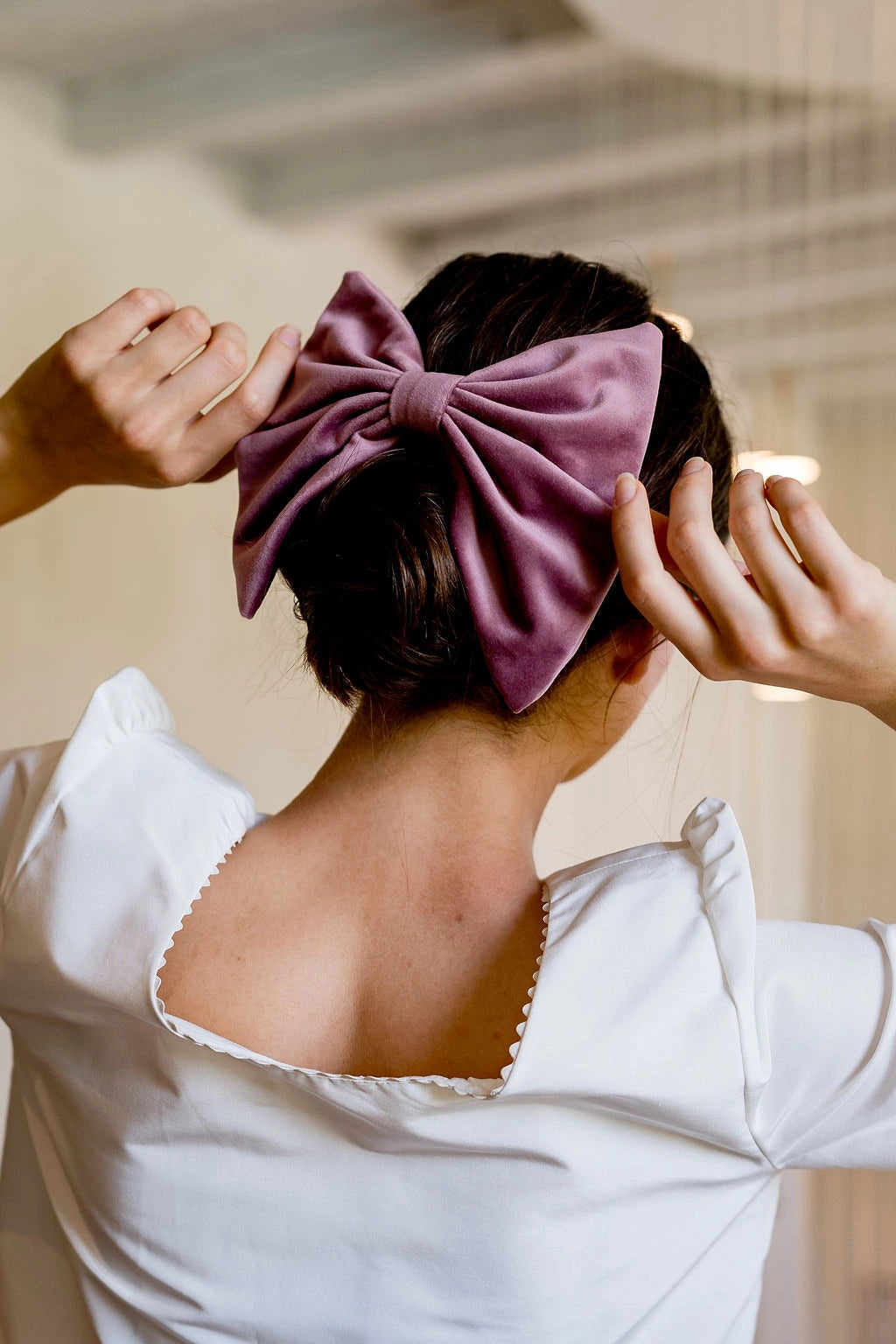 Jeune femme se tenant de dos avec un haut blanc ayant dans les cheveux un noeud en velours de couleur figue monté sur barrette et piqué au dessus de son chignon. Créé par Bellemond.