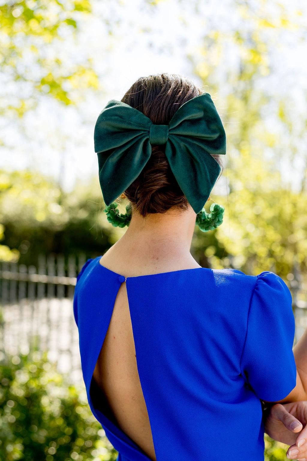 Jeune femme se tenant de dos dans une robe bleue roi avec dans les cheveux une barrette en forme de noeud en velours vert sapin. Créé par Bellemond. 