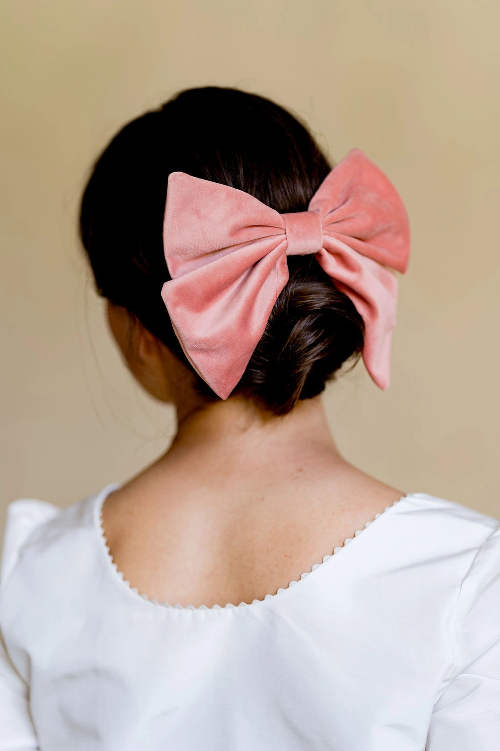 Jeune femme se tenant de dos avec un haut blanc ayant dans les cheveux un noeud de couleur vieux rose en matière de velours monté sur une barrette et glissée au dessus de son chignon. Créé par Bellemond. 