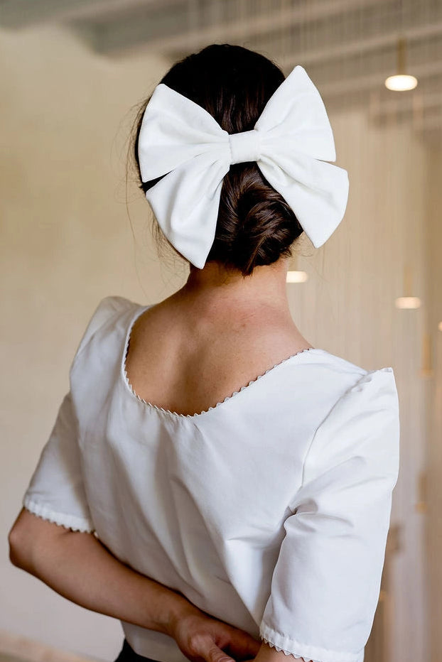 Femme se tenant de dos avec un haut blanc ayant dans les cheveux un gros noeud en velours de type barrette. Créé par Bellemond.