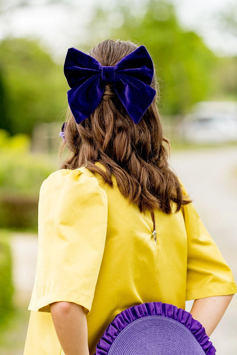 Jeune femme se tenant de dos habillée d'une robe jaune et portant dans les cheveux une barrette avec un noeud en velours de couleur violet. Créé par Bellemond. 